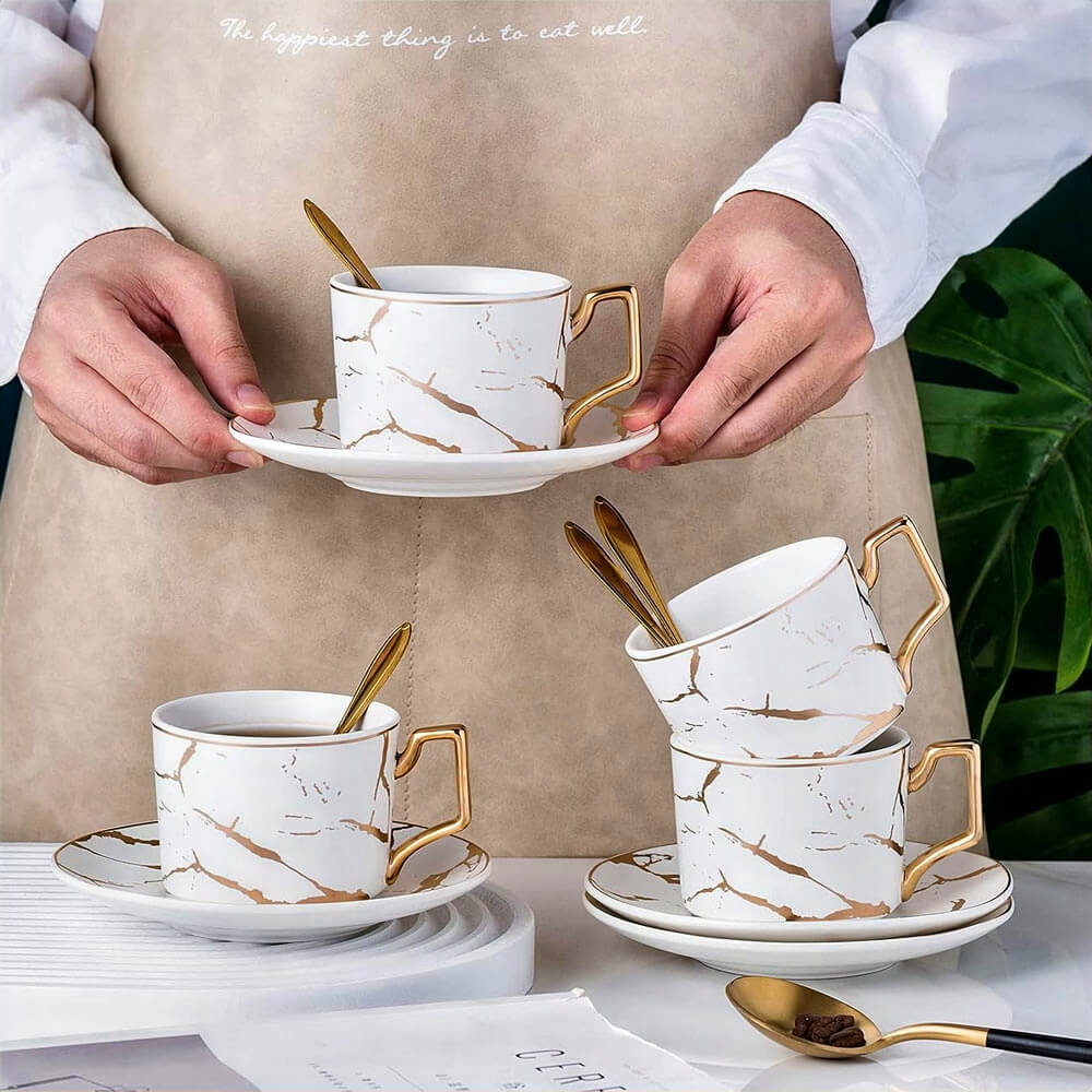 A stylish set of teacups and saucers with a marble design, gold accents, and matching spoons. A person holds one cup, highlighting its elegant craftsmanship.