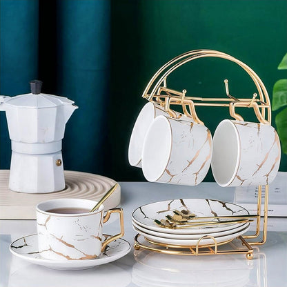 A sophisticated teacups saucers set displayed elegantly on a gold-tone rack. The teacups feature a marble design with gold accents, paired with matching saucers and golden spoons. A filled teacup is set beside the rack, and a white coffee maker complements the scene.