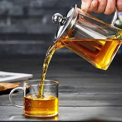 A hand pouring tea from a clear glass teapot with a stainless steel infuser into a matching glass cup on a dark wooden table.