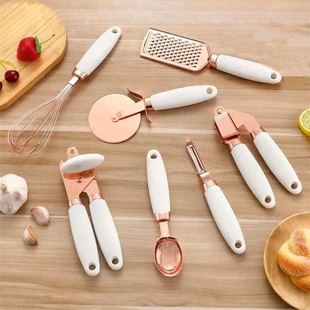 A beautifully designed tool set for the kitchen featuring rose gold accents and white ergonomic handles, including a whisk, grater, pizza cutter, garlic press, can opener, vegetable peeler, and ice cream scoop, elegantly arranged on a wooden countertop alongside fresh ingredients for a stylish culinary setup.