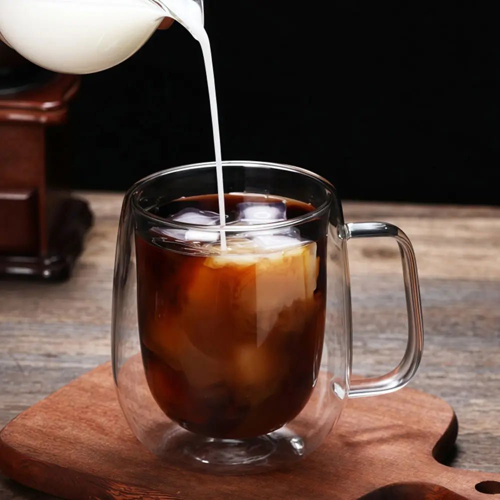 Glass mug with iced coffee being topped with milk on a wooden surface.