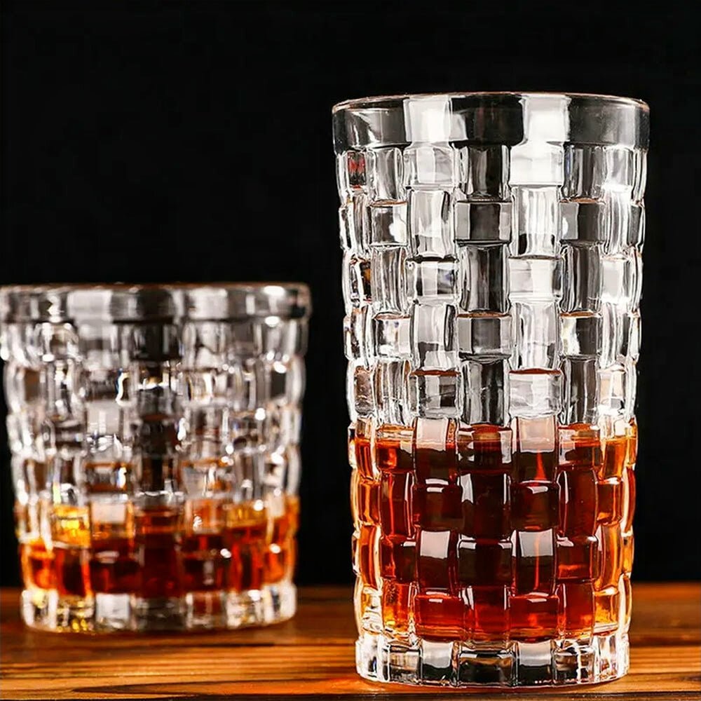 Two vintage highball glasses with a geometric lattice pattern are shown. One is taller and partially filled with an amber liquid, while the other, shorter glass is similarly filled. They are set on a wooden surface against a dark background, highlighting the intricate texture and clarity of the glass.