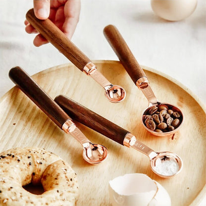 A set of vintage measuring spoons with copper-plated heads and dark walnut handles displayed on a wooden tray. One spoon is being held, while others are filled with ingredients like coffee beans and flour, surrounded by a bagel, eggshell, and a cozy kitchen setting.