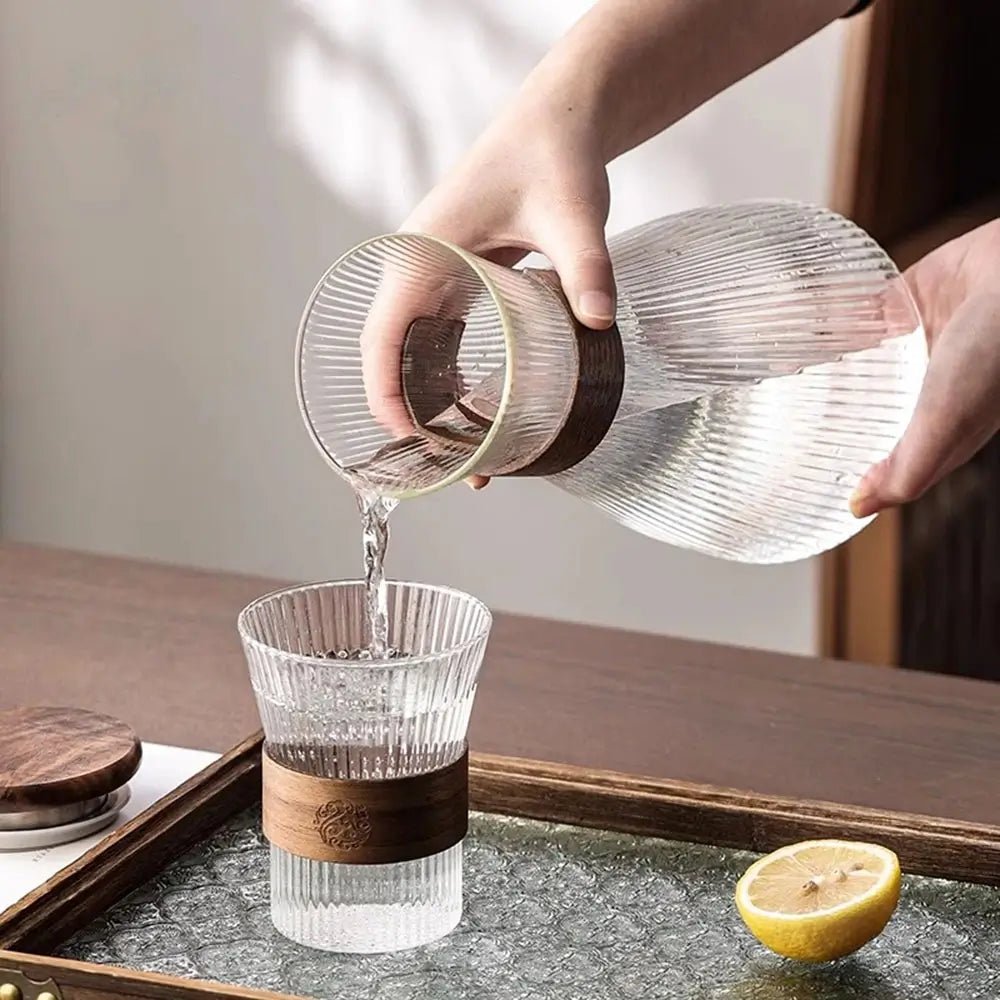 Hand pouring water from ribbed glass pitcher with wooden band into matching ribbed glass on tray with half lemon.