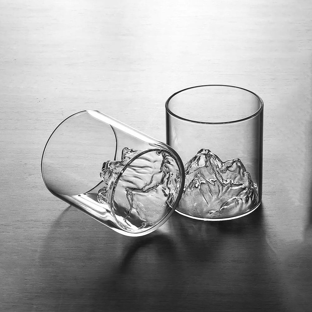 Two whiskey glasses with mountains in the bottom are displayed, featuring a unique and intricate mountain peak design sculpted into the base. One glass stands upright while the other lies on its side, emphasizing the clear glass construction and showcasing the detailed craftsmanship of the mountain-inspired interior.