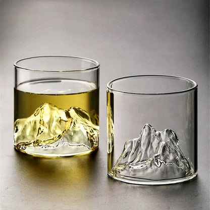 Two whiskey glasses with mountains are displayed, each featuring a sculpted mountain peak design at the base. One glass is filled with a golden liquid, showcasing how the beverage accentuates the intricate details of the mountain design, while the other glass remains empty, highlighting the clarity and craftsmanship of the glass. 