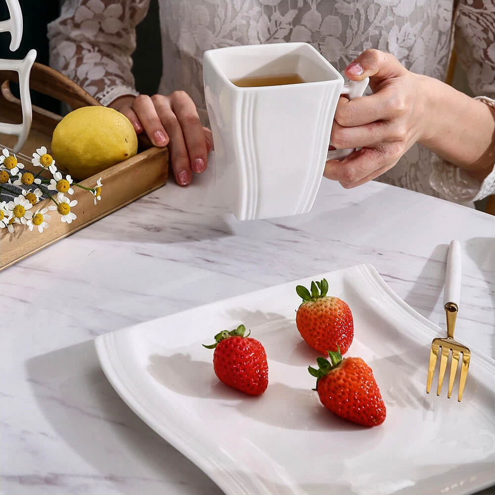 Woman holding a white porcelain coffee mug with tea, next to a plate of strawberries and a gold fork.